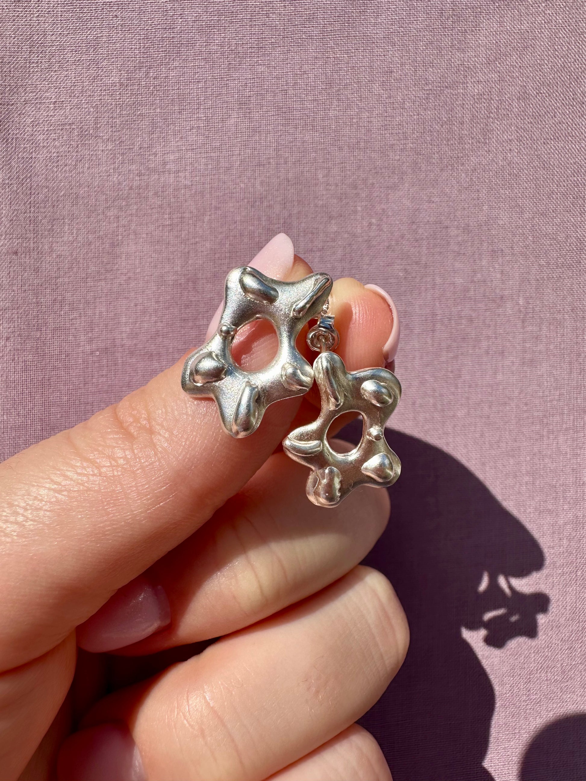 Silver star-shaped earrings held between fingers against a pink background