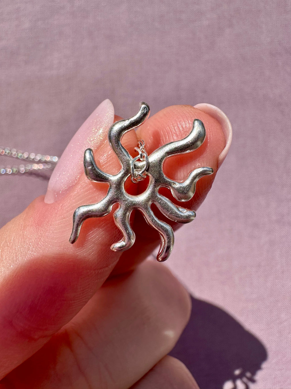 Silver sun pendant held between fingers against a pink background