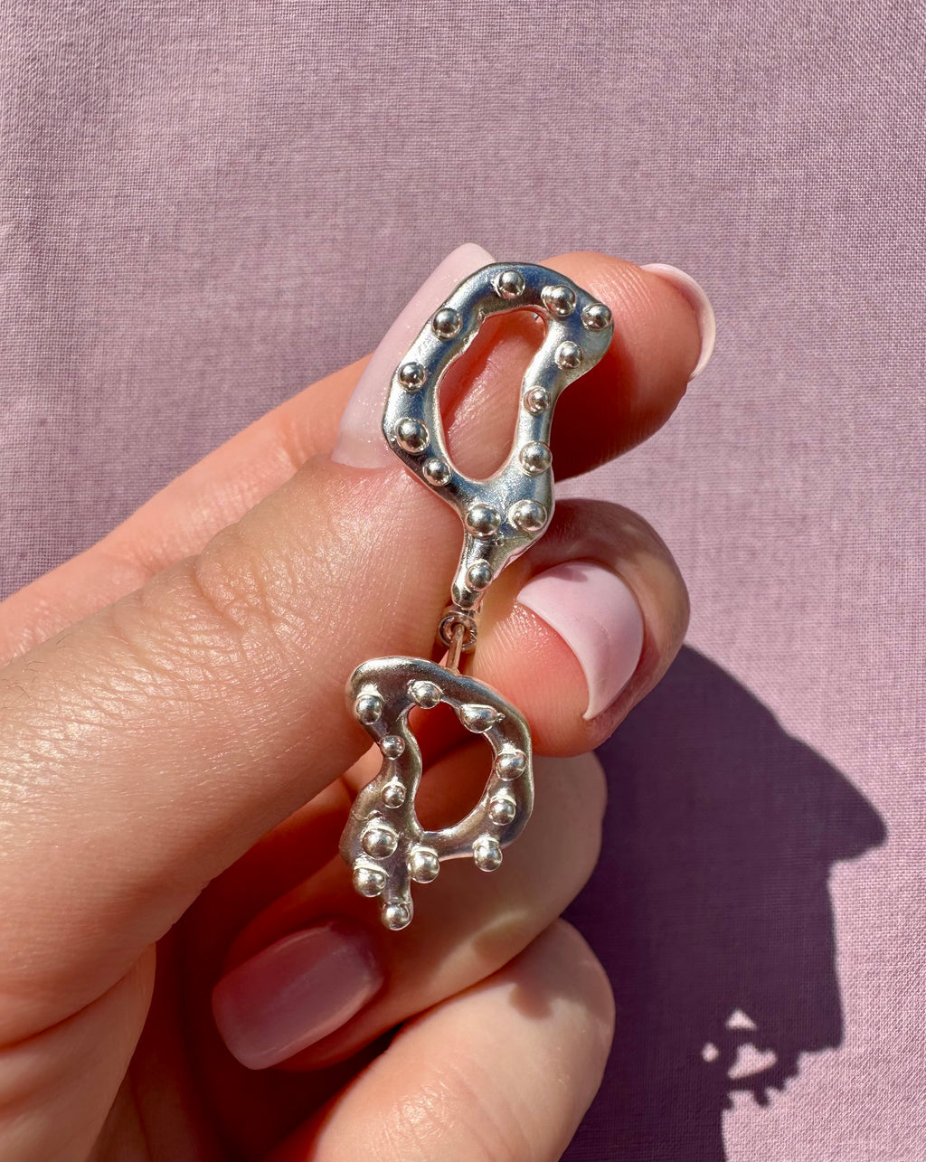 Silver earrings held by a hand against a pink background