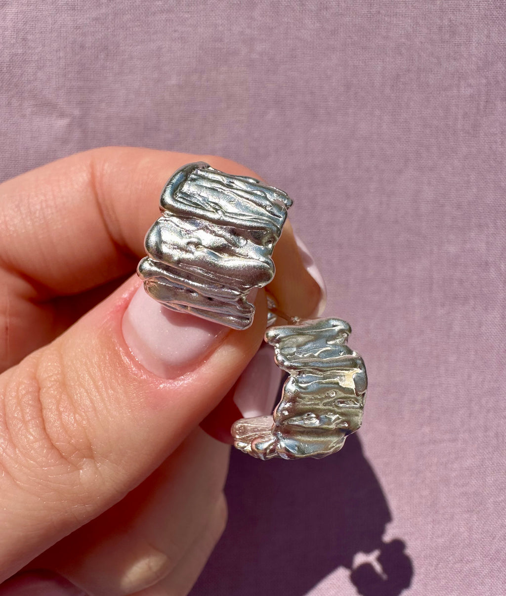 Silver earring with textured design held by a hand against a pink background