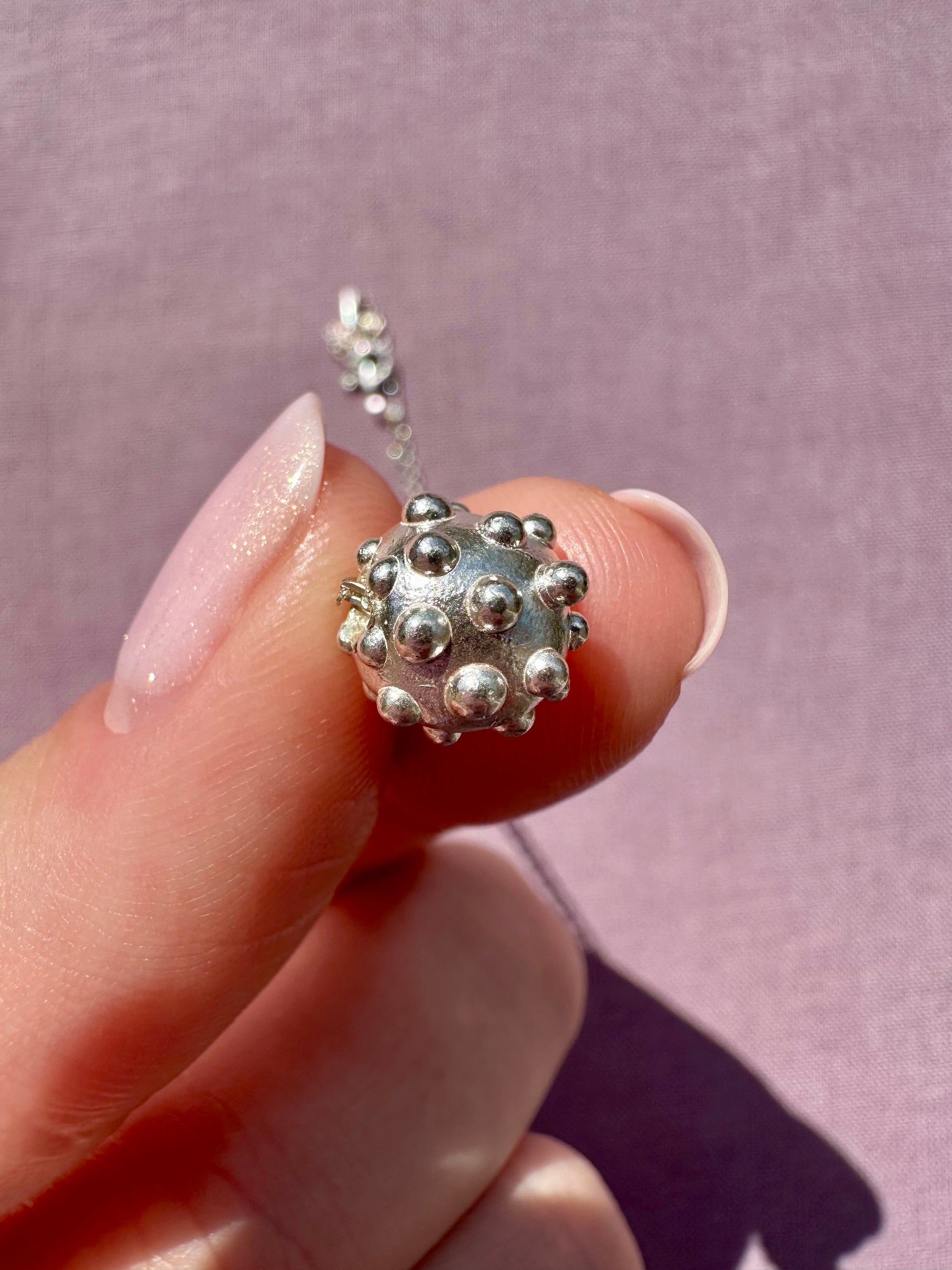 Silver textured bead on a chain held between fingers against a pink background