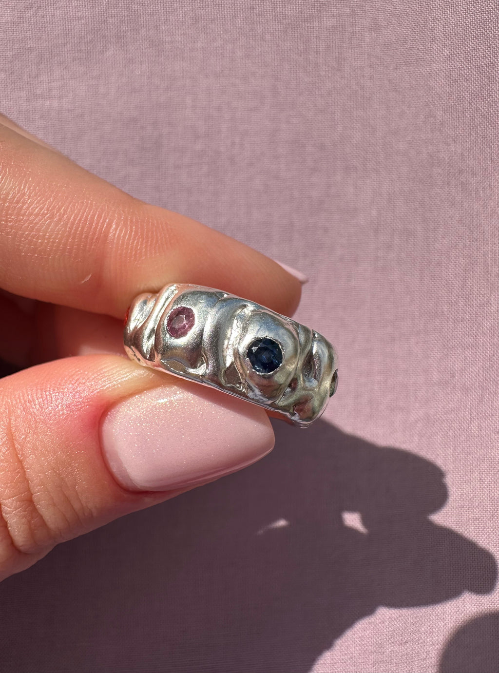 Silver ring with gemstones held by a hand on a pink background