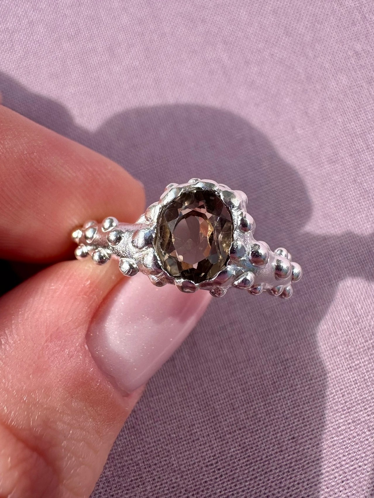 Silver ring with a central gemstone held by a hand against a light purple background