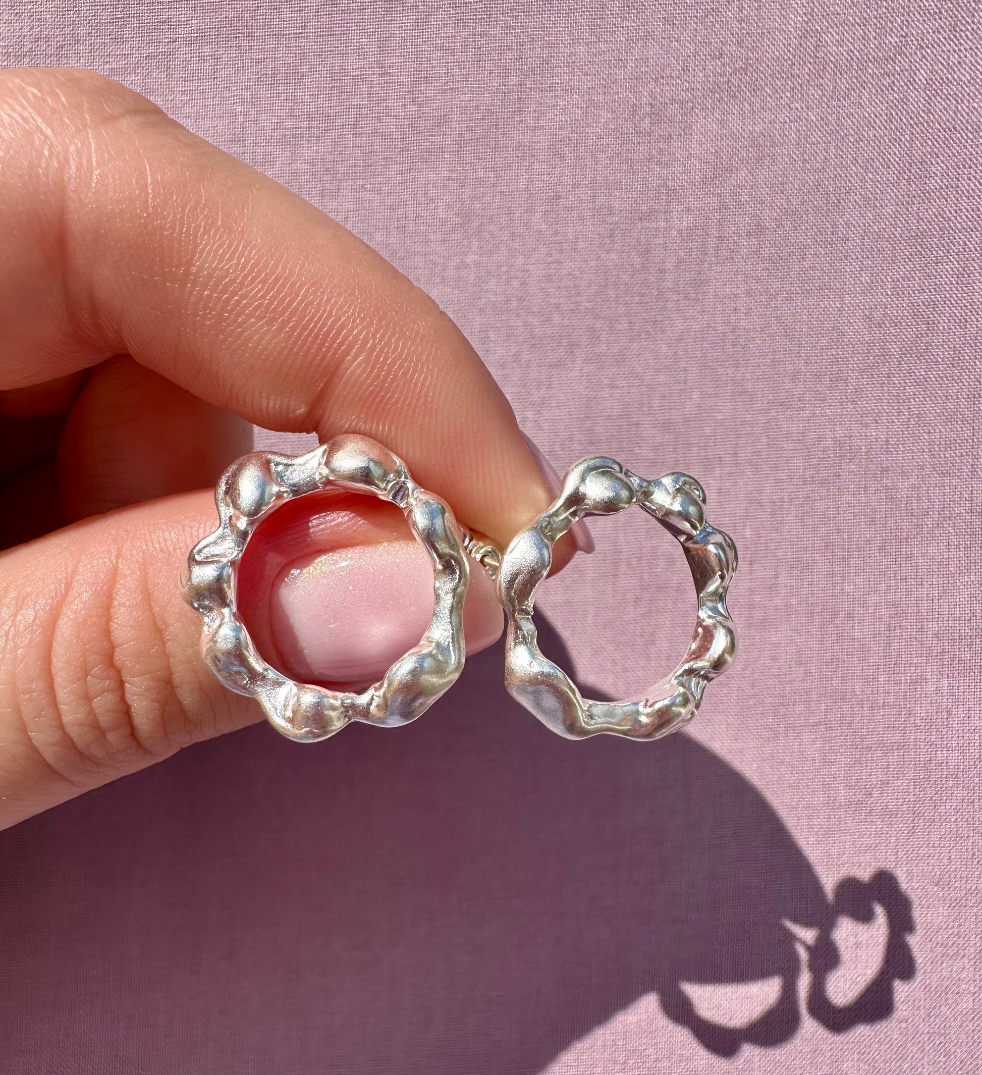 Silver earrings held between fingers against a pink background