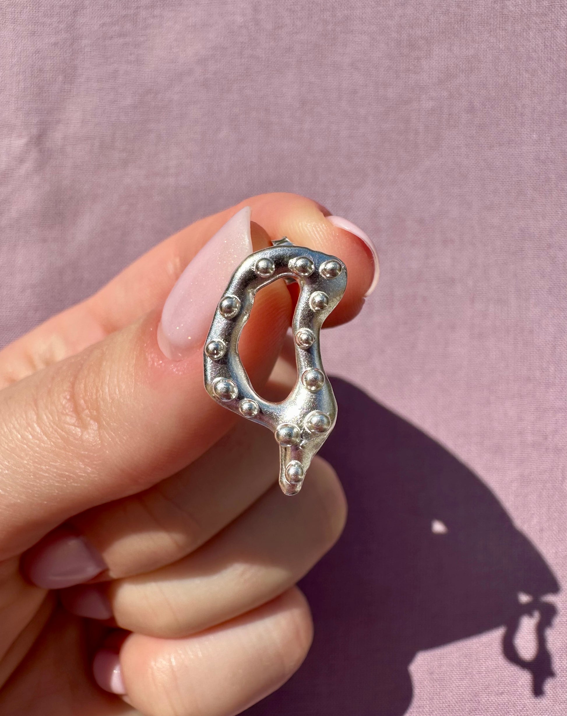 Silver earring with bead detailing held by a hand against a pink background