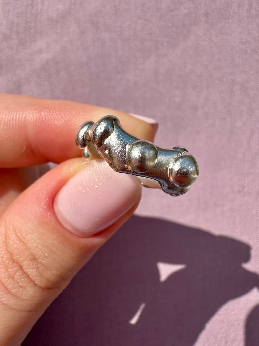 Silver ring with three pebble-like designs held by a hand against a pink background