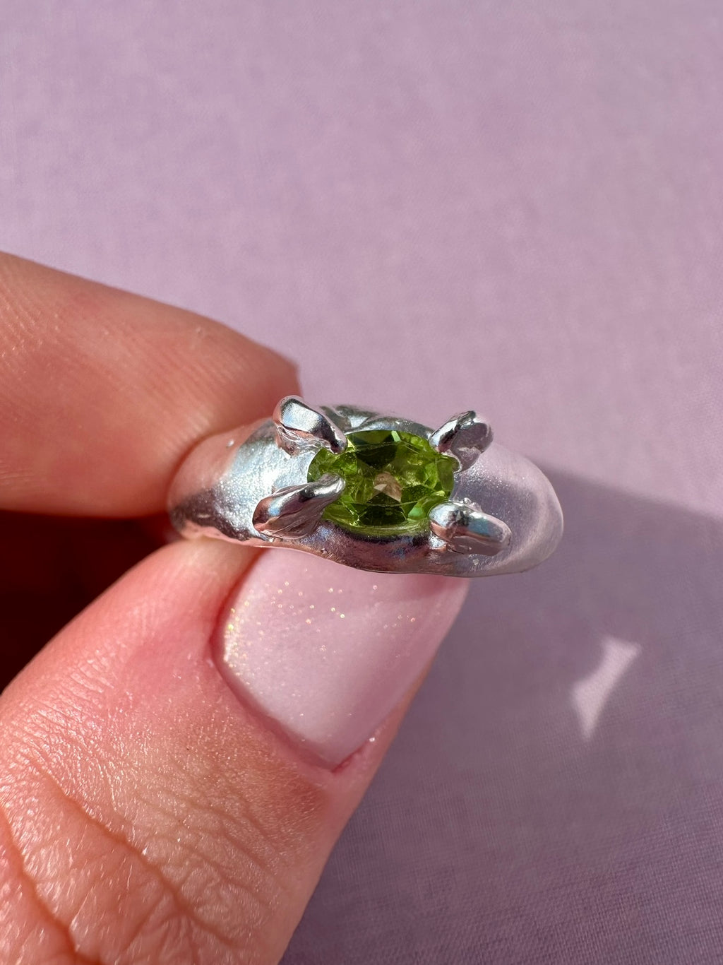 Silver ring with a green gemstone held between fingers on a light purple background